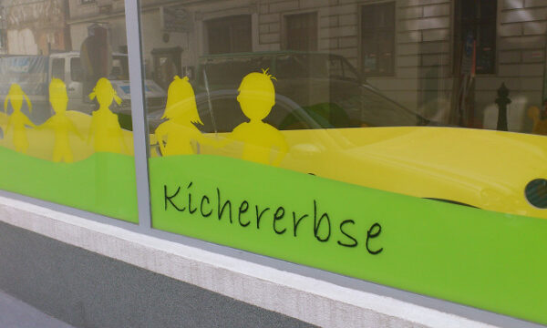 Fenster mit gelben Kindersilhouetten und dem Wort "Kichererbse" auf grünen und gelben Tafeln, mit Gebäuden und einem Auto, das sich im Glas spiegelt.