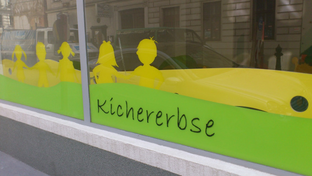 Fenster mit gelben Kindersilhouetten und dem Wort "Kichererbse" auf grünen und gelben Tafeln, mit Gebäuden und einem Auto, das sich im Glas spiegelt.