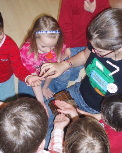 Eine Gruppe kleiner Kinder versammelt sich um sie und streckt ihre Hände aus, um etwas zu berühren oder zu betrachten, das ein Mädchen mit Brille und schwarzem T-Shirt in der Hand hält.