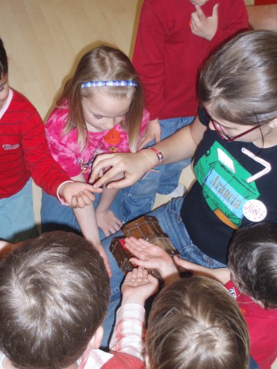 Eine Gruppe kleiner Kinder versammelt sich um sie und streckt ihre Hände aus, um etwas zu berühren oder zu betrachten, das ein Mädchen mit Brille und schwarzem T-Shirt in der Hand hält.