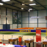 Indoor-Sportanlage mit einem kleinen Fußballfeld mit Absperrungen, Kickertischen im Vordergrund und Bannern an den Wänden, die für "Wien sport & fun" werben.