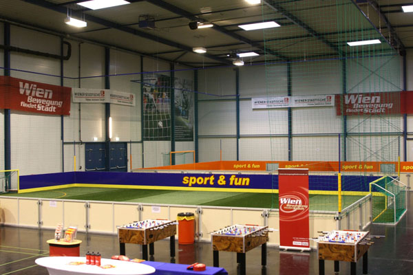 Indoor-Sportanlage mit einem kleinen Fußballfeld mit Absperrungen, Kickertischen im Vordergrund und Bannern an den Wänden, die für "Wien sport & fun" werben.