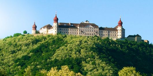 Ein großes historisches Gebäude mit roten Türmen thront auf einem grünen, baumbestandenen Hügel unter strahlend blauem Himmel.