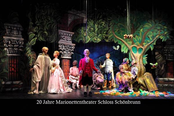 Eine Marionettentheateraufführung mit kunstvoll gekleideten Puppen auf einer farbenfrohen Bühne mit Waldmotiven. Text: „20 Jahre Marionettentheater Schloss Schönbrunn.“.