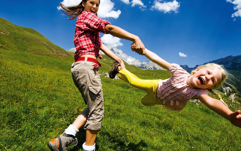Zwei Kinder spielen draußen auf einer Wiese, wobei das eine das andere an den Armen herumwirft, unter einem blauen Himmel mit vereinzelten Wolken.