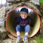 Ein kleiner Junge mit blauer Mütze und Turnschuhen sitzt in einem runden Spielplatztunnel aus Stein und Metall und lächelt in die Kamera.