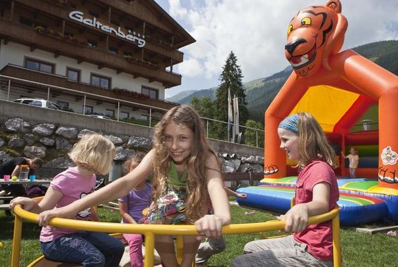 Vier Kinder spielen auf einem Karussell vor einem Gebäude, mit einer orangefarbenen, tigerförmigen Hüpfburg im Hintergrund auf einer Wiese.