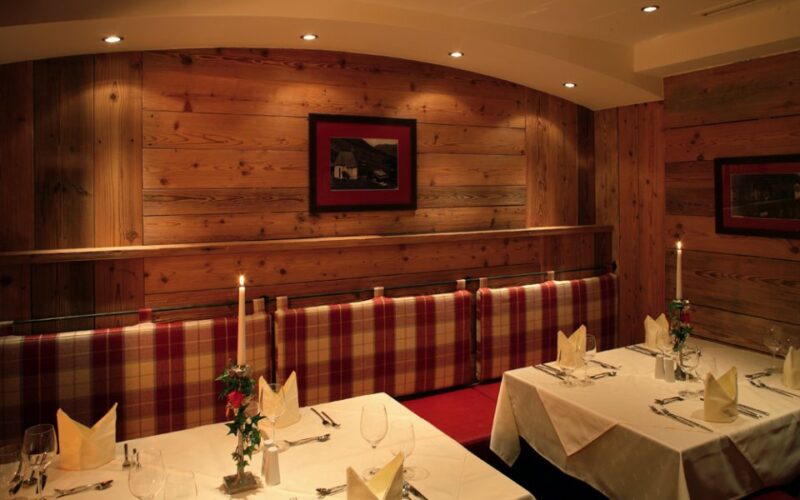 Ein gemütlicher Restaurantbereich mit Holzwänden, gepolsterten Sitzen, gedeckten Tischen mit weißen Tüchern, Kerzen und gerahmten Bildern an den Wänden.