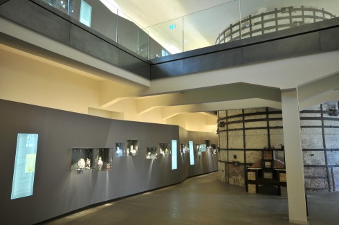 Ein modernes Museumsinterieur mit Glasvitrinen an der Wand, in denen verschiedene Artefakte ausgestellt sind, und einer freiliegenden, runden Backsteinstruktur im Hintergrund.