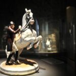 Eine Porzellanfigur eines Mannes in Uniform, der neben einem sich aufbäumenden weißen Pferd steht, ausgestellt auf einem runden Sockel in einem schwach beleuchteten Ausstellungsraum.