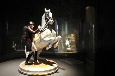 Eine Porzellanfigur eines Mannes in Uniform, der neben einem sich aufbäumenden weißen Pferd steht, ausgestellt auf einem runden Sockel in einem schwach beleuchteten Ausstellungsraum.