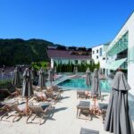 Außenpoolbereich mit Liegestühlen und geschlossenen Sonnenschirmen neben einem modernen Gebäude, vor der Kulisse der Berge und unter einem blauen Himmel.