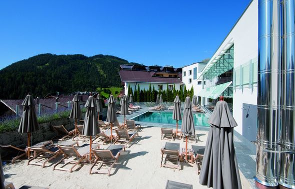 Außenpoolbereich mit Liegestühlen und geschlossenen Sonnenschirmen neben einem modernen Gebäude, vor der Kulisse der Berge und unter einem blauen Himmel.