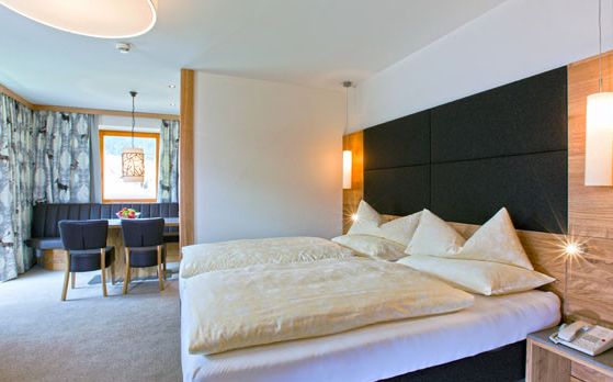 Helles Hotelzimmer mit einem Doppelbett, gepolstertem Kopfteil, Tisch und Stühlen am Fenster und moderner Beleuchtung.