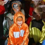 Kinder in farbenfrohen Kostümen sitzen zusammen, wobei ein Kind in einem orangefarbenen Outfit und mit Gesichtsbemalung in der Mitte lächelt.