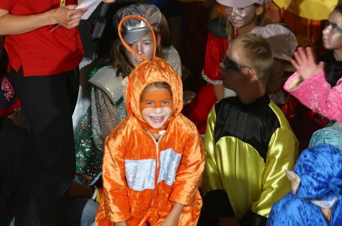 Kinder in farbenfrohen Kostümen sitzen zusammen, wobei ein Kind in einem orangefarbenen Outfit und mit Gesichtsbemalung in der Mitte lächelt.