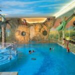 Hallenbad mit tropischem Dschungel-Thema, mit Rutschen, bemalten Wänden, Palmen-Dekorationen und Kindern, die im Wasser spielen.