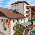 Ein traditionelles Alpenhotel mit weißen Wänden, Holzbalkonen, Blumenkästen und einer Terrasse mit grünen Sonnenschirmen vor einem teilweise bewölkten Himmel.