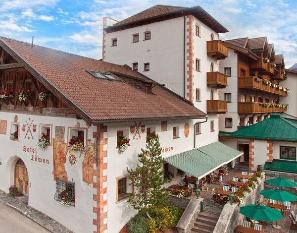 Ein traditionelles Alpenhotel mit weißen Wänden, Holzbalkonen, Blumenkästen und einer Terrasse mit grünen Sonnenschirmen vor einem teilweise bewölkten Himmel.