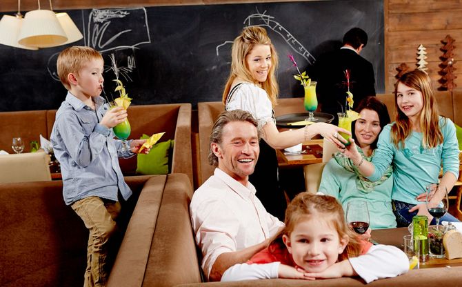 Eine Familie sitzt zusammen in einem Restaurant, lächelt und genießt die Getränke, während ein Kellner einem der Erwachsenen einen Cocktail reicht.