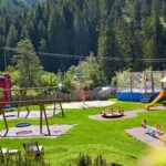 Spielplatz im Freien mit Rutschen, Schaukeln, einem Karussell, einem Trampolin, einer aufblasbaren Rutsche und Wippen auf grünem Gras, umgeben von Bäumen und Bergen im Hintergrund.