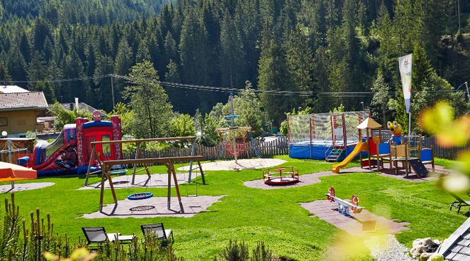 Spielplatz im Freien mit Rutschen, Schaukeln, einem Karussell, einem Trampolin, einer aufblasbaren Rutsche und Wippen auf grünem Gras, umgeben von Bäumen und Bergen im Hintergrund.