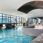 Moderne Schwimmhalle mit großen Fenstern, einer geschwungenen Metallskulptur, Steinstufen und Sitzgelegenheiten. Der Raum ist von natürlichem Licht durchflutet; draußen sind Bäume und Berge zu sehen.