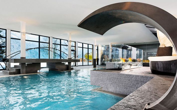 Moderne Schwimmhalle mit großen Fenstern, einer geschwungenen Metallskulptur, Steinstufen und Sitzgelegenheiten. Der Raum ist von natürlichem Licht durchflutet; draußen sind Bäume und Berge zu sehen.