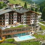 Ein großes Gebirgsresort mit Wasserrutsche, Freibad, Spielplatz und grünen Hügeln inmitten eines Alpendorfs.