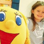 Ein junges Mädchen und ein Junge stehen lächelnd neben einem großen gelben Plüschtier mit Gesicht in einem Haus.