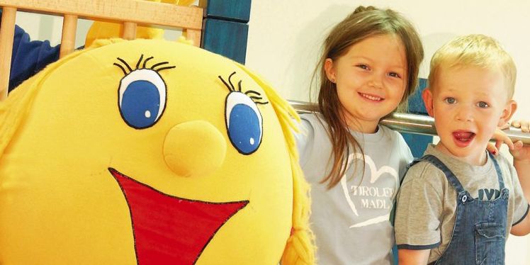 Ein junges Mädchen und ein Junge stehen lächelnd neben einem großen gelben Plüschtier mit Gesicht in einem Haus.