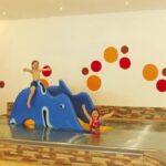 Drei Kinder spielen in einem kleinen Hallenbad mit bunten, tierförmigen Strukturen und orangefarbenen, roten Wanddekorationen.