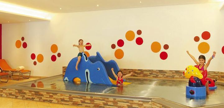 Drei Kinder spielen in einem kleinen Hallenbad mit bunten, tierförmigen Strukturen und orangefarbenen, roten Wanddekorationen.