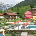 Alpine Ferienanlage mit Gebäuden im Chalet-Stil, Außenpool, roten Sonnenschirmen, gelber Wasserrutsche, Spielplatz und Berglandschaft im Hintergrund.