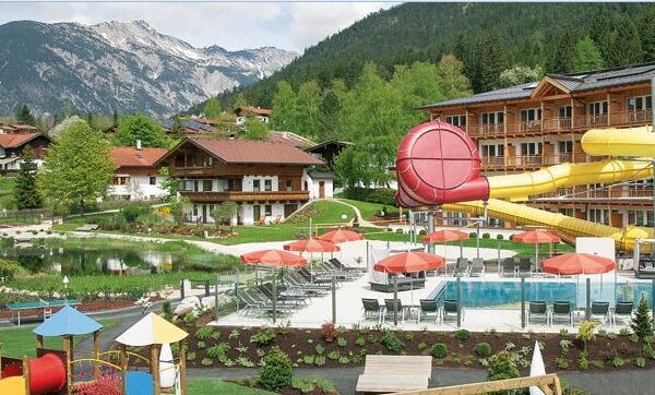 Alpine Ferienanlage mit Gebäuden im Chalet-Stil, Außenpool, roten Sonnenschirmen, gelber Wasserrutsche, Spielplatz und Berglandschaft im Hintergrund.