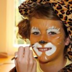 Eine Person in einem Leopardenkostüm hat ihr Gesicht mit weißer, schwarzer und orangefarbener Farbe so bemalt, dass es wie ein Löwe aussieht.