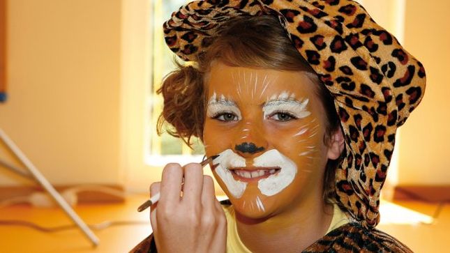 Eine Person in einem Leopardenkostüm hat ihr Gesicht mit weißer, schwarzer und orangefarbener Farbe so bemalt, dass es wie ein Löwe aussieht.