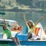 Vier Personen, zwei Erwachsene und zwei Kinder, sitzen auf einem Tretboot auf einem See, lächeln und winken mit erhobenen Armen. Im Hintergrund sind ein Segelboot und Grünflächen zu sehen.