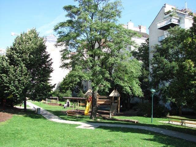 Ein kleiner Spielplatz mit einer Rutsche und einem Klettergerüst befindet sich inmitten von Bäumen und Gras, mit Wohnhäusern im Hintergrund.