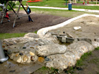 Ein Spielbereich für Kinder im Freien mit einem Wasser- und Sandbereich mit Steinen und einer kleinen Pumpe, der an einen Spielplatz mit Schaukeln angrenzt.