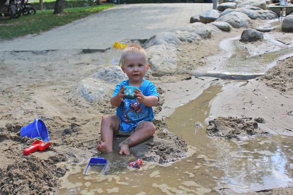 Ein Kleinkind in einem blauen Hemd sitzt in einer sandigen, schlammigen Gegend an einem kleinen Bach und spielt mit einem Eimer und einer Schaufel Spielzeug.