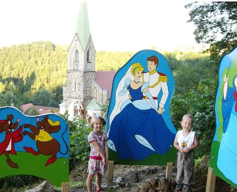 Zwei kleine Kinder stehen vor farbenfrohen Ausschnitten, die Aschenputtel und andere Märchenfiguren darstellen, mit einer Kirche und Grünpflanzen im Hintergrund.