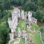 Luftaufnahme einer steinernen Burgruine inmitten eines dichten grünen Waldes, mit einem Fluss in der Nähe und einem kleinen Haus im Hintergrund.
