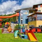 Spielplatz im Freien an einem Hotel mit einer roten Wasserrutsche, Spielgeräten, einer Spielzeugeisenbahn und einer Kängurustatue auf grünem Gras unter einem teilweise bewölkten Himmel.