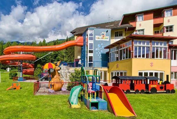 Spielplatz im Freien an einem Hotel mit einer roten Wasserrutsche, Spielgeräten, einer Spielzeugeisenbahn und einer Kängurustatue auf grünem Gras unter einem teilweise bewölkten Himmel.