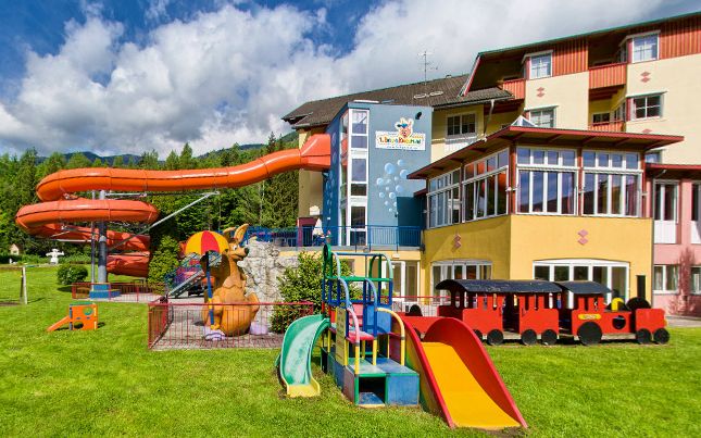 Spielplatz im Freien an einem Hotel mit einer roten Wasserrutsche, Spielgeräten, einer Spielzeugeisenbahn und einer Kängurustatue auf grünem Gras unter einem teilweise bewölkten Himmel.
