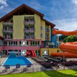 Das Hotel verfügt über Balkone mit Blick auf einen Außenpool, Liegestühle und eine orangefarbene Wasserrutsche, die sich an einem sonnigen Tag in den Poolbereich hineinwindet.