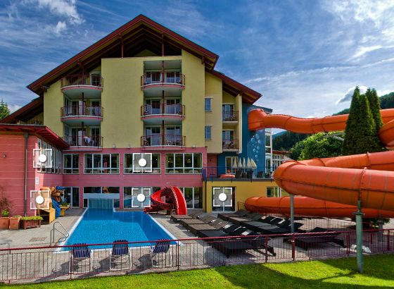Das Hotel verfügt über Balkone mit Blick auf einen Außenpool, Liegestühle und eine orangefarbene Wasserrutsche, die sich an einem sonnigen Tag in den Poolbereich hineinwindet.
