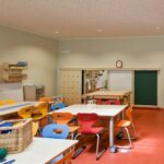 Klassenzimmer mit bunten Stühlen und Tischen, Regalen mit Material und einem Bastelkorb auf dem Tisch. Der Raum hat einen roten Teppich und neutrale Wände.