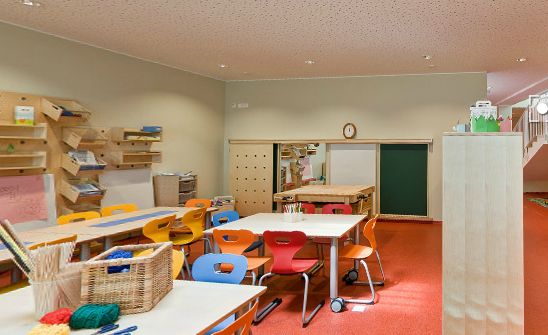 Klassenzimmer mit bunten Stühlen und Tischen, Regalen mit Material und einem Bastelkorb auf dem Tisch. Der Raum hat einen roten Teppich und neutrale Wände.